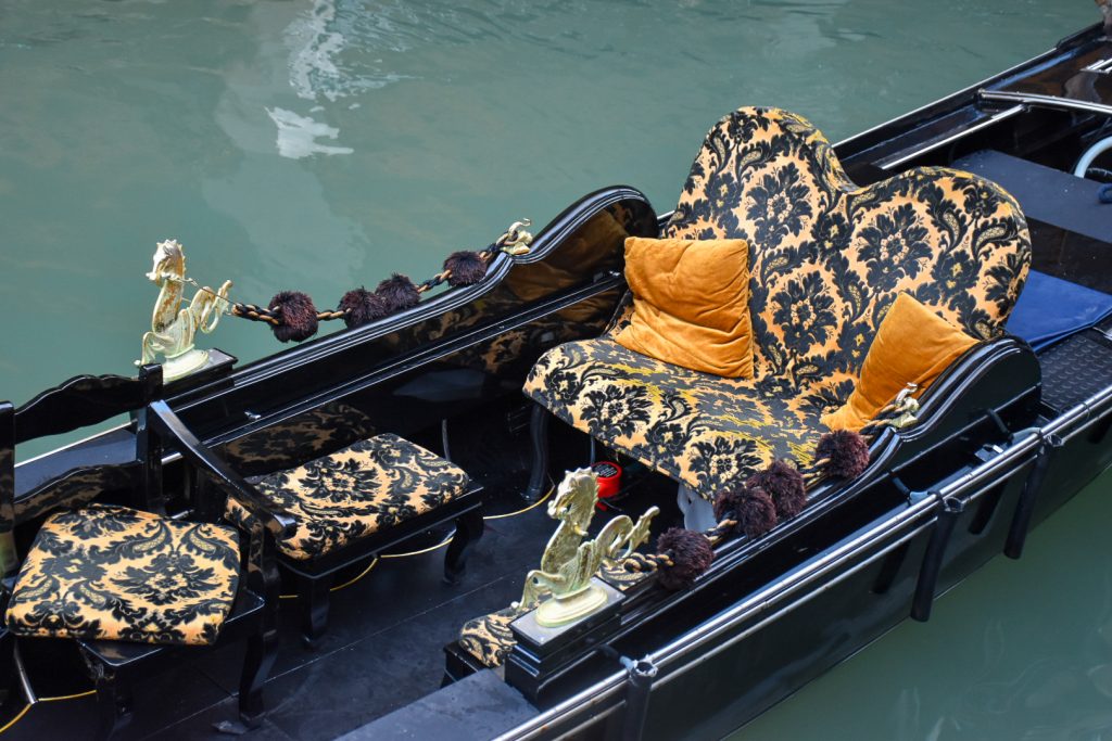 Looking down into the body of a gondola boat. The gondola is black lacquer and has a gold and black seat with orange cushions. Still waters either side