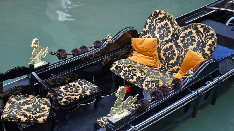 Looking down into the body of a gondola boat. The gondola is black lacquer and has a gold and black seat with orange cushions. Still waters either side