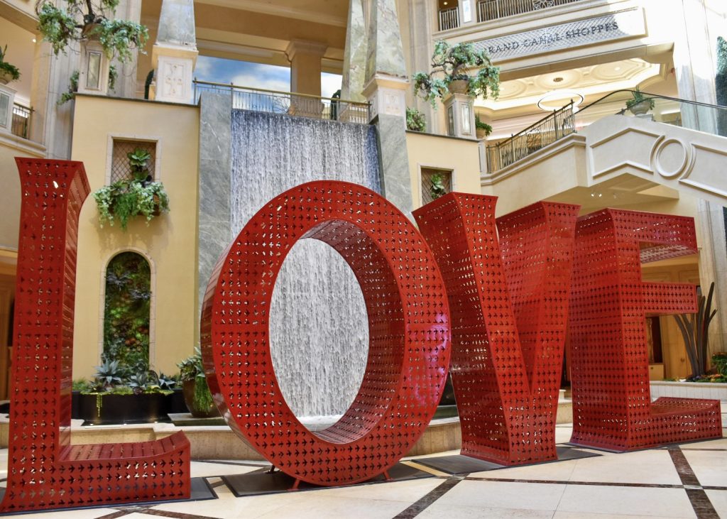Large red metal letters spelling LOVE sitting on a floor in a shopping centre. There is a water wall behind the word and an escalator up to the upper floor of the shopping centre