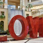 Large red metal letters spelling LOVE sitting on a floor in a shopping centre. There is a water wall behind the word and an escalator up to the upper floor of the shopping centre