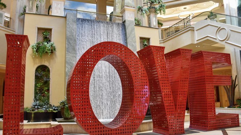 Large red metal letters spelling LOVE sitting on a floor in a shopping centre. There is a water wall behind the word and an escalator up to the upper floor of the shopping centre