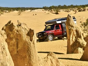 Yellow desert of sand with large rock formations in the foreground and more rocks towards the far side of the frame, along with some green bushland and pale blue sky. A red camper van with silver roof is parked on the sand in the middle of the photo behind one of the pinnacles