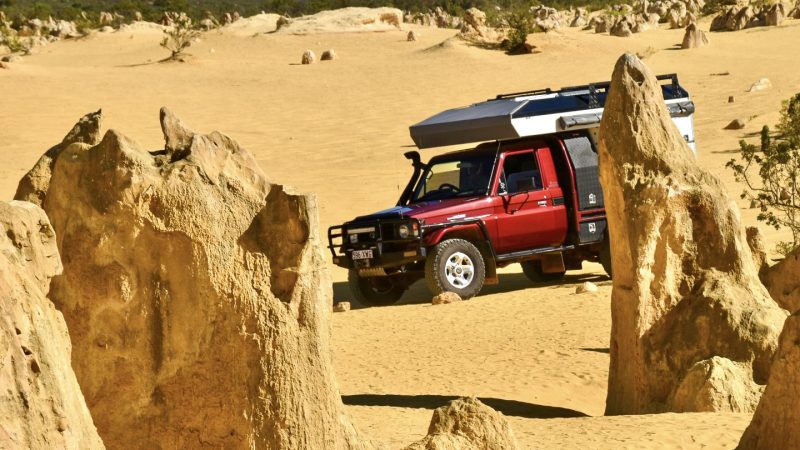 Yellow desert of sand with large rock formations in the foreground and more rocks towards the far side of the frame, along with some green bushland and pale blue sky. A red camper van with silver roof is parked on the sand in the middle of the photo behind one of the pinnacles