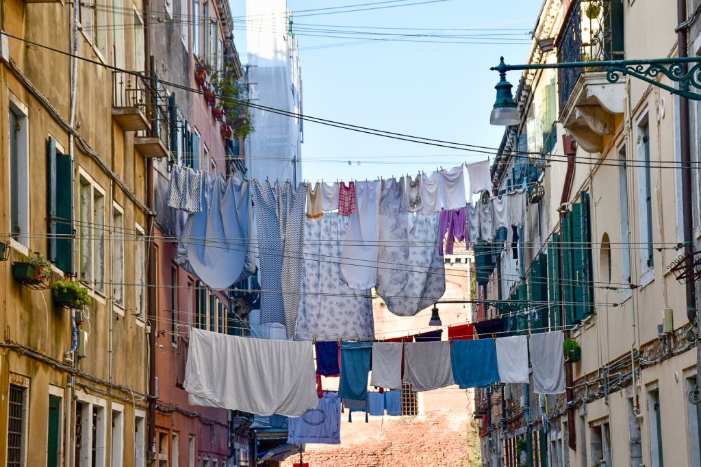 Lines of washing strung between two tall buildings with shutters and iron balconies. Sheets, towels and clothes hang in-between