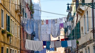 Lines of washing strung between two tall buildings with shutters and iron balconies. Sheets, towels and clothes hang in-between