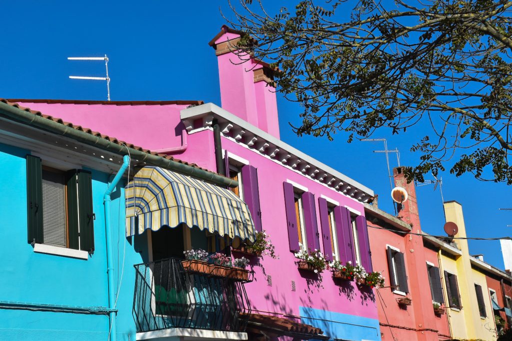 Deep blue sky at the top. In the bottom two thirds of the photo is a row of colourful houses painted - turquoise, pink with purple shutters, orange, yellow. The turquoise house has a striped sun shade over its balcony with pots of flowers. Tree branches and leaves feature in the top right of photo