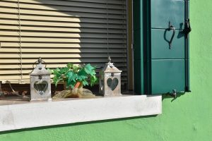 A bright green wall shows along the bottom of the photo and up the right hand side. There is a window ledge to the left of the photo featuring two silver lanterns and a green plant, wooden slatted blinds and a dark green window shutter to the right