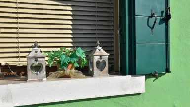 A bright green wall shows along the bottom of the photo and up the right hand side. There is a window ledge to the left of the photo featuring two silver lanterns and a green plant, wooden slatted blinds and a dark green window shutter to the right