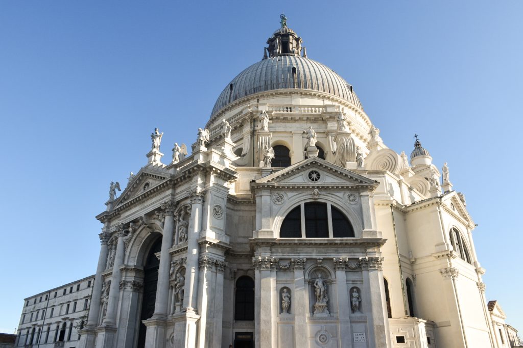 Pale blue clear sky above. Large white marble Baroque church in the front and centre of photo