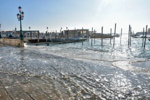 Scene of the water front with the the sunshine in the background. Water is flooded onto the pavement areas. Gondola boats and wooden posts on the water
