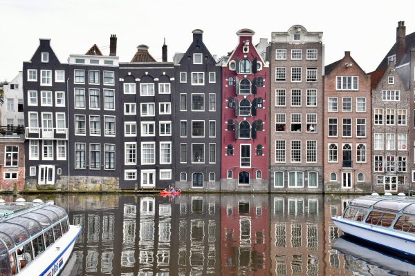 Photo taken across a canal, looking at a row of multicoloured Dutch houses which are leaning and uneven. Two canal boats wait on the water on left and right hand side