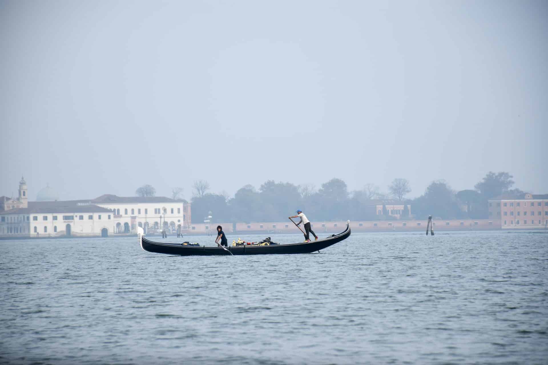 Misty scene across water to white buildings and trees on the horizon. A long black gondola boat is making its way across the centre of the image with two people rowing