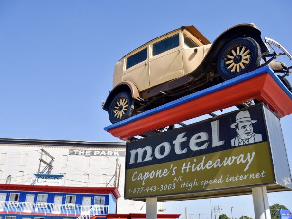A sign for a motel called Capone's Hideaway appears from the bottom right of the frame, Above the sign is an old-fashioned cream car (1930s) mounted on a platform - looks like a gangster car. Bottom left shows the top of a white building with blue and red bedrooms. Blue sky above