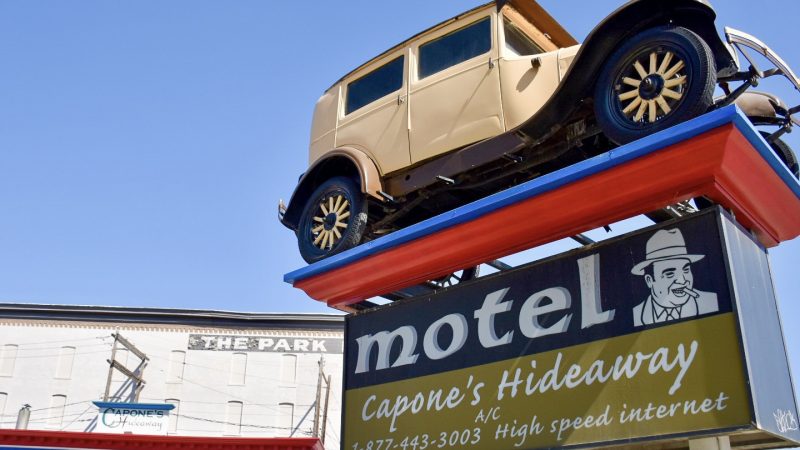 A sign for a motel called Capone's Hideaway appears from the bottom right of the frame, Above the sign is an old-fashioned cream car (1930s) mounted on a platform - looks like a gangster car. Bottom left shows the top of a white building with blue and red bedrooms. Blue sky above
