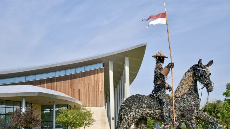 Modern building made of wood with a sky jump shaped roof in the bottom middle section of the frame. Blue sky with cover of grey cloud behind. Bottom left features a sculpture of a man on horseback. The man is wearing a Canadian Mountie hat and holding a red and white flag on a pole