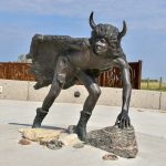 Large brass sculpture in the foreground. It is of a character with a cape made of animal skins and horns on the head. The man is leaning over as it looking for something in the distance. Blue sky, grass and a curved barrier behind