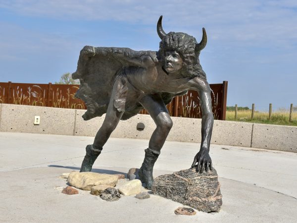 Large brass sculpture in the foreground. It is of a character with a cape made of animal skins and horns on the head. The man is leaning over as it looking for something in the distance. Blue sky, grass and a curved barrier behind