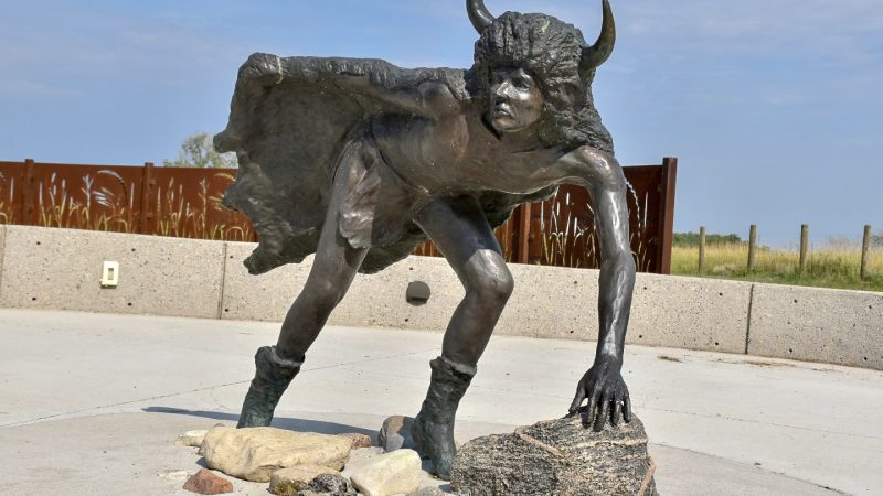 Large brass sculpture in the foreground. It is of a character with a cape made of animal skins and horns on the head. The man is leaning over as it looking for something in the distance. Blue sky, grass and a curved barrier behind