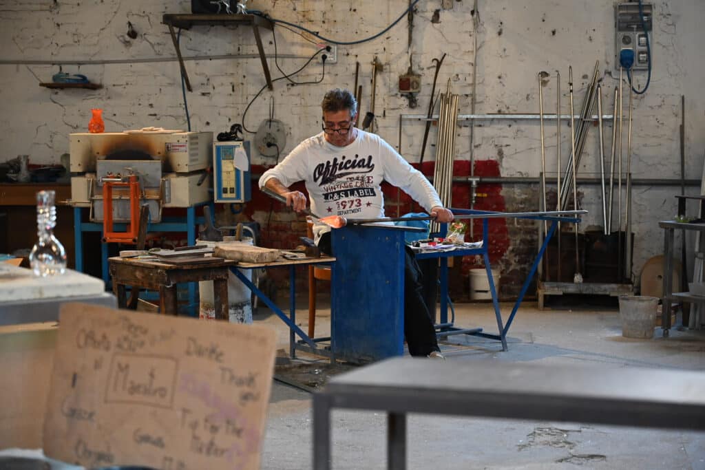 Glass maestro at the OMG workshop on Burano shaping a glowing gather of molten glass on a long pipe, with the furnace behind him and a handwritten “Thank you Maestro” sign in the foreground.