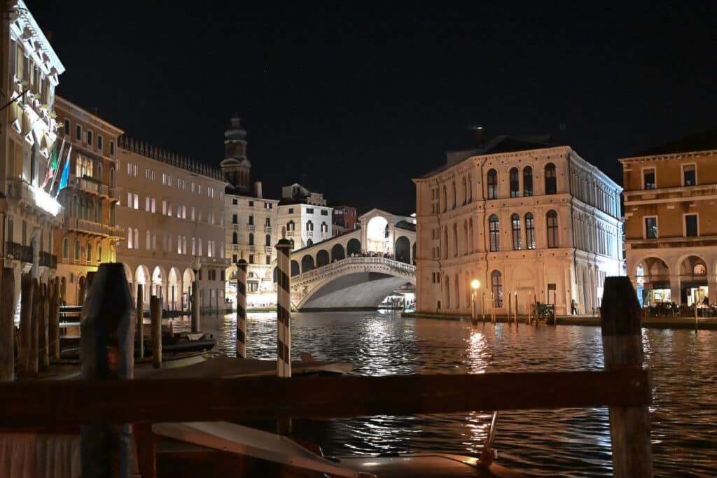 Night view of the Rialto Bridge over the Grand Canal, lit white against a dark sky, with palazzi on both banks, striped mooring poles in the foreground and lights reflecting on the water.