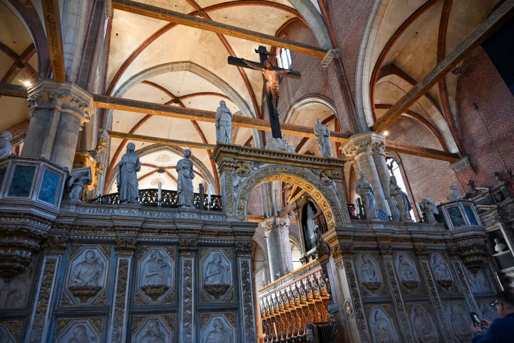 Interior of the Frari in Venice looking at the marble choir screen, with a wooden crucifix above the central arch, stone apostles along the top, carved relief panels below, and Gothic brick vaults overhead.