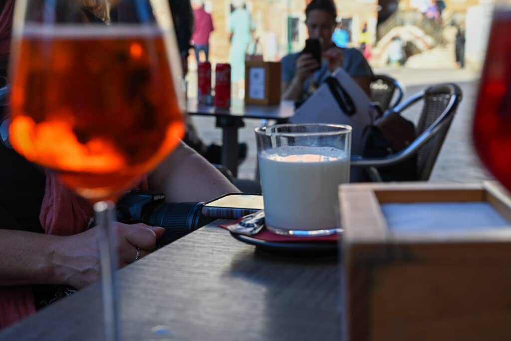 Outdoor café table in Campo San Barnaba, Venice, with a blurred Aperol spritz in the foreground, a glass of milky coffee in focus, a Nikon camera and phone on the table, and people sitting and walking in the square behind.