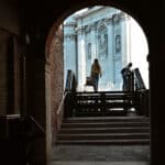 Dark brick archway in Venice framing a sunlit view of Santa Maria della Salute, with two people climbing the bridge steps and a wall plaque reading Campo de la Salute.