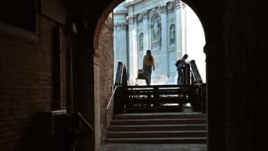 Dark brick archway in Venice framing a sunlit view of Santa Maria della Salute, with two people climbing the bridge steps and a wall plaque reading Campo de la Salute.