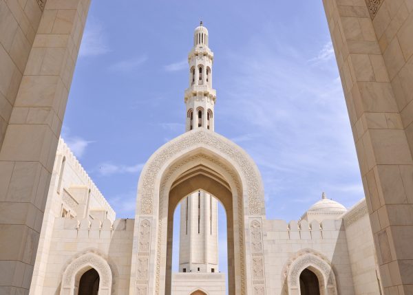 Carved white marble arches and minaret of mosque. Pale blue sky behind. Arabic designs