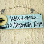Pale yellow painted wall with a wooden sign hanging by a piece of wire on two nails hammered into the wall. The wooden sign is painted turquoise and says 'Relax...Unwind... It's Margarita Time'