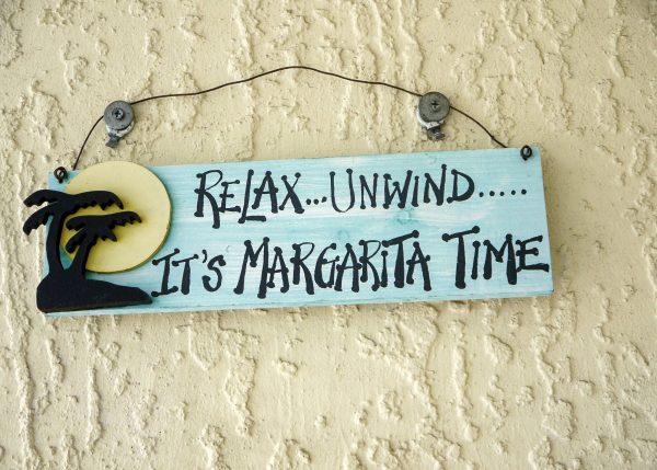 Pale yellow painted wall with a wooden sign hanging by a piece of wire on two nails hammered into the wall. The wooden sign is painted turquoise and says 'Relax...Unwind... It's Margarita Time'