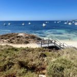 Blue sky over a blue and turquoise sea, with white motor boats moored in the water.. Grassland n the foreground and wooden staircase leading down to a white beach