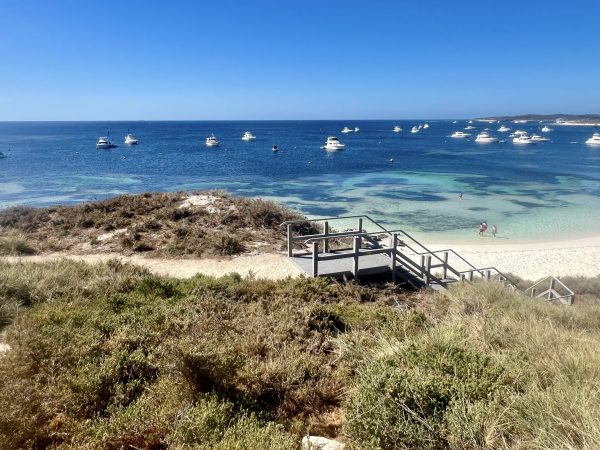 Blue sky over a blue and turquoise sea, with white motor boats moored in the water.. Grassland n the foreground and wooden staircase leading down to a white beach
