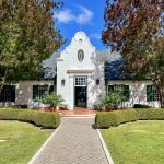 A white building stretches across the photo with green shuttered windows - two windows to either side of a tall doorway in the centre. Trees either side and blue sky above. There is a pathway leading from the middle front towards the building. It's a very symmetrical photo