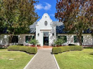 A white building stretches across the photo with green shuttered windows - two windows to either side of a tall doorway in the centre. Trees either side and blue sky above. There is a pathway leading from the middle front towards the building. It's a very symmetrical photo