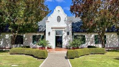 A white building stretches across the photo with green shuttered windows - two windows to either side of a tall doorway in the centre. Trees either side and blue sky above. There is a pathway leading from the middle front towards the building. It's a very symmetrical photo