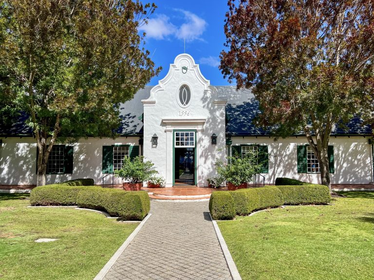 A white building stretches across the photo with green shuttered windows - two windows to either side of a tall doorway in the centre. Trees either side and blue sky above. There is a pathway leading from the middle front towards the building. It's a very symmetrical photo