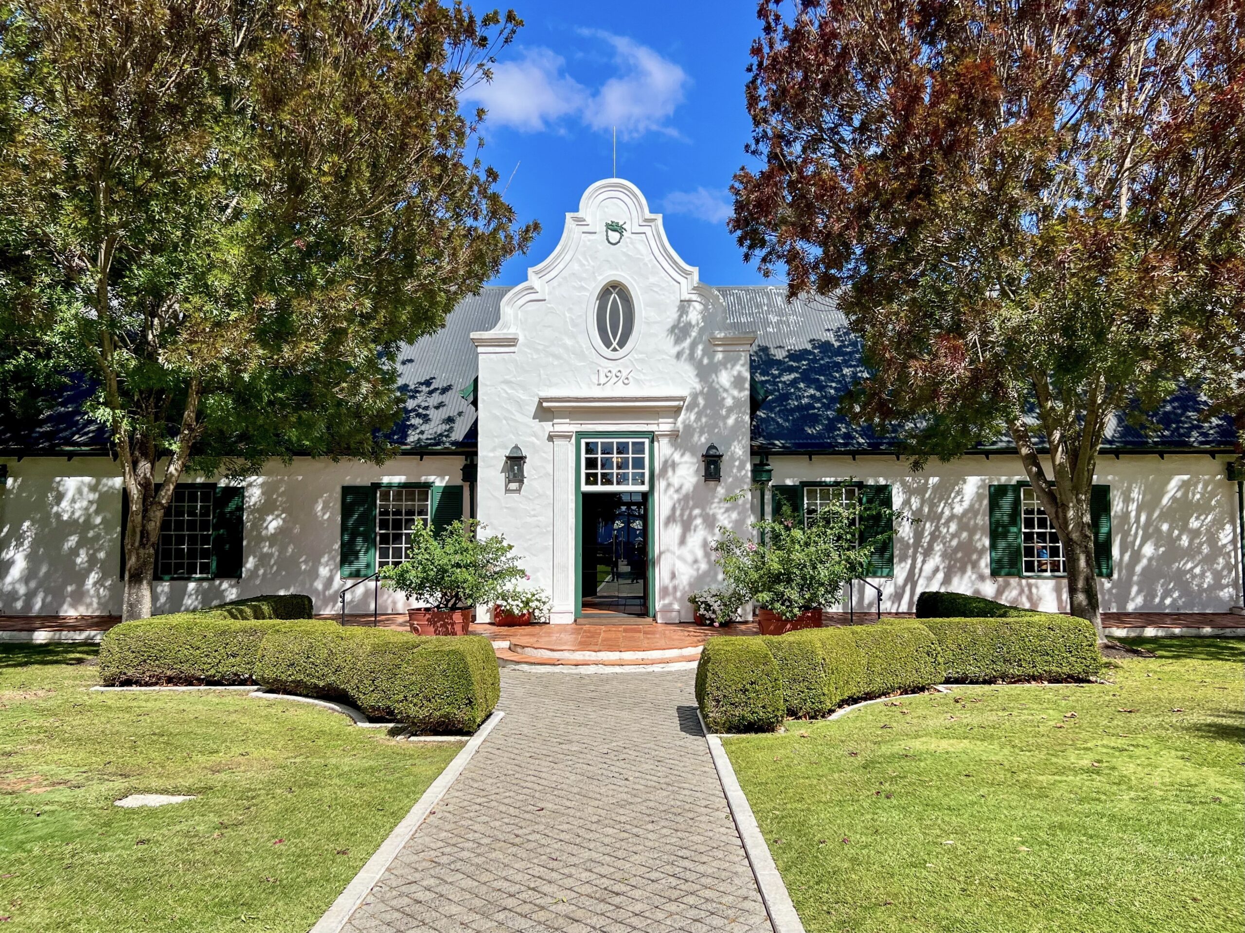 A white building stretches across the photo with green shuttered windows - two windows to either side of a tall doorway in the centre. Trees either side and blue sky above. There is a pathway leading from the middle front towards the building. It's a very symmetrical photo