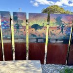 Sculpture made of a series of 6 panels stretching across the middle of the photo. The bottom of the panels are made of dark wood and the top of the panels feature Australian indigenous art work of figures, animals and birds