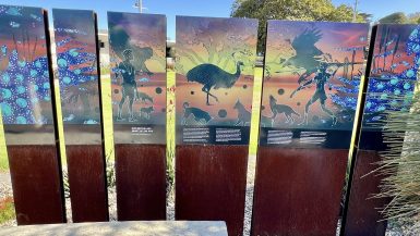 Sculpture made of a series of 6 panels stretching across the middle of the photo. The bottom of the panels are made of dark wood and the top of the panels feature Australian indigenous art work of figures, animals and birds