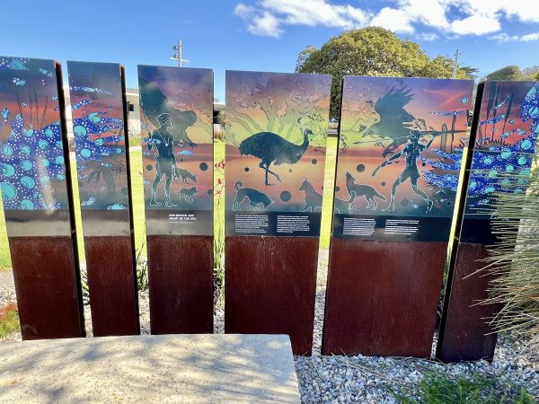 Sculpture made of a series of 6 panels stretching across the middle of the photo. The bottom of the panels are made of dark wood and the top of the panels feature Australian indigenous art work of figures, animals and birds