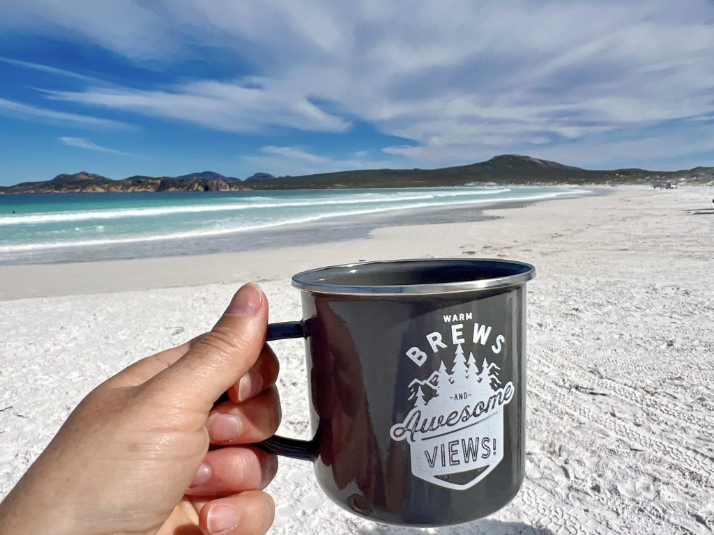 A hand in the foreground holds out a black metal cup which says 'Warm Brews and Awesome Views!'. Behind is a sweeping powder white beach and turquoise waves across the bay. On the horizon are some hills with a cloudy blue sky above.