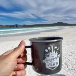 A hand in the foreground holds out a black metal cup which says 'Warm Brews and Awesome Views!'. Behind is a sweeping powder white beach and turquoise waves across the bay. On the horizon are some hills with a cloudy blue sky above.