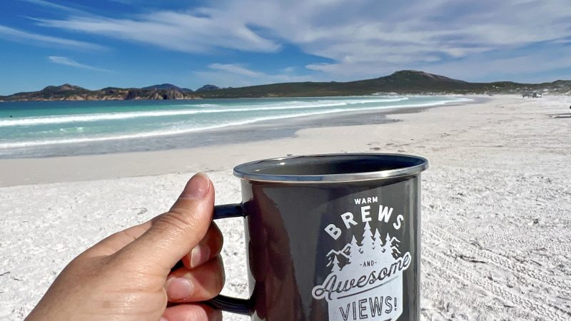 A hand in the foreground holds out a black metal cup which says 'Warm Brews and Awesome Views!'. Behind is a sweeping powder white beach and turquoise waves across the bay. On the horizon are some hills with a cloudy blue sky above.
