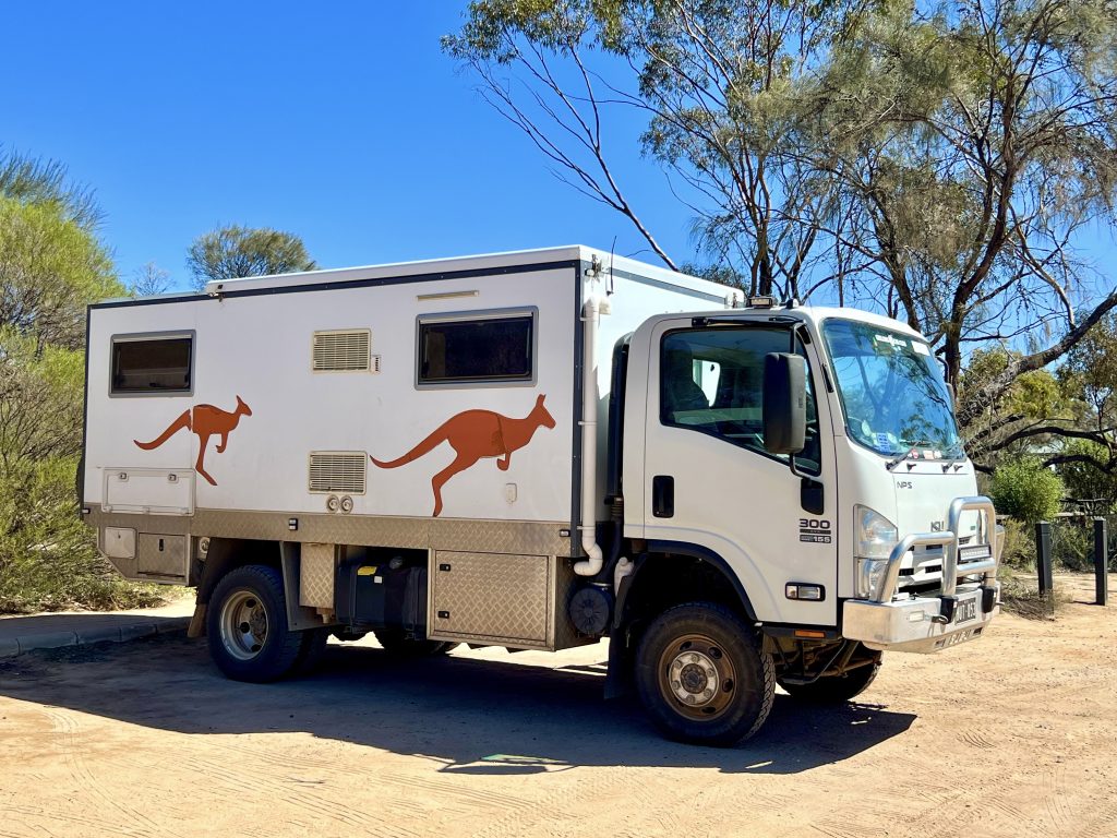 Small white lorry van with 2 brown jumping kangaroos printed on the side. Parked on sandy ground with blue sky and green shrubs and trees behind