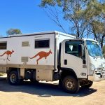 Small white lorry van with 2 brown jumping kangaroos printed on the side. Parked on sandy ground with blue sky and green shrubs and trees behind