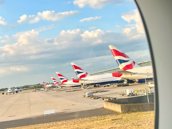 Pale blue sky with fluffy white clouds in top half. Tarmac ground on the bottom. Row of 4 white aircraft lined up on the right hand side. The aircraft have red, white and blue Union Jack branding on the tail fins, the British Airways logo