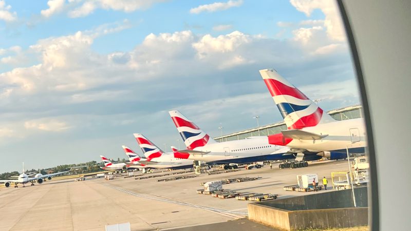 Pale blue sky with fluffy white clouds in top half. Tarmac ground on the bottom. Row of 4 white aircraft lined up on the right hand side. The aircraft have red, white and blue Union Jack branding on the tail fins, the British Airways logo