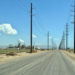 Blue sky at the top of frame. A dusty straight road leads from bottom front of photo towards the horizon. There are tall telegraph poles supporting wires on either side of the road. To the left of the road is a factory, otherwise the landscape looks barren