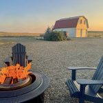 Sundown shot of a lit fire pit in the bottom right of photo with two great chairs around the fire pit. On the horizon is an expanse of grassland and a white barn with a red roof illuminated in the sunset